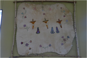 Scene of the crossroads embroidered on a moose hide and mounted on a square wooden frame with rope. We also see embroidered flowers. The whole is installed on the wall. Picture in color.