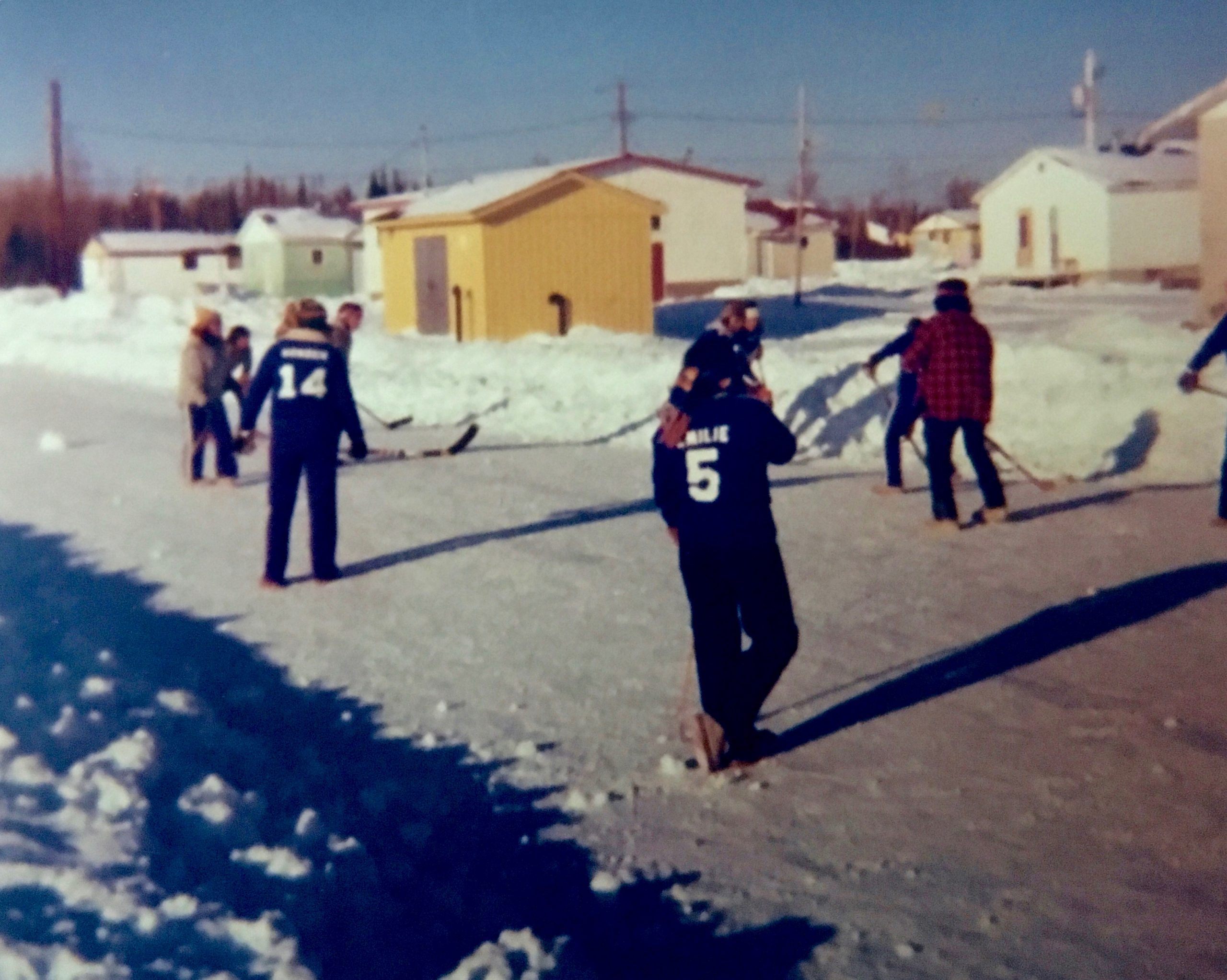 Anicinabek youth playing field hockey in the streets of the community. The road is snowy and snow lines the driveways. We can see some houses around. Picture in color.