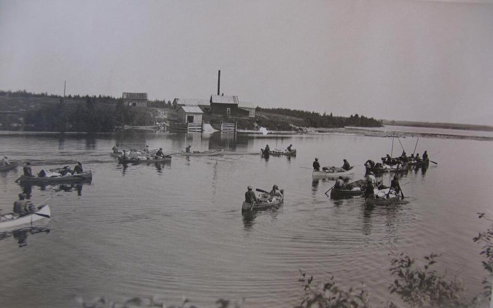 Families arriving by canoe at the summer gathering. We see a dozen canoes with people on board and luggage. On the shore, we see some small houses. Black and white picture.