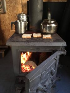 Tea and toast are heating on a wood stove with the door open, allowing the fire to be seen. Three pots of tea and three slices of bread are cooking on top of the stove. Picture in color.