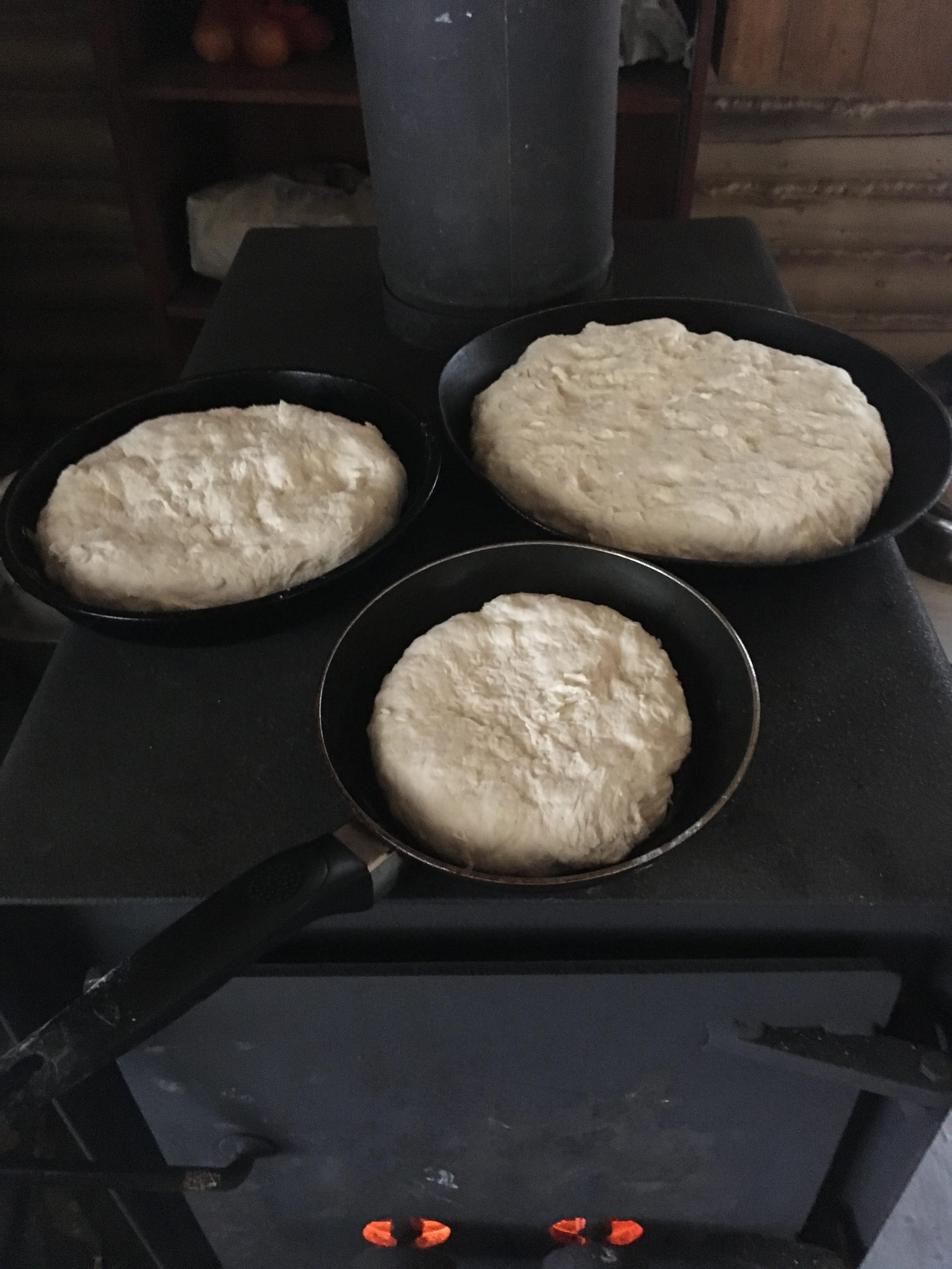 Three bannocks are cooking in pans placed directly on a wood pole. Picture in color.