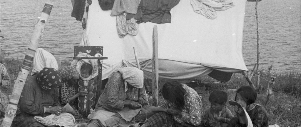 Anicinabek women sit on the shore of Lac Simon. They are sewing. Children are watching them. A tent with cloth on it is in the background. The picture dates from the middle of the 20th century. Black and white picture.