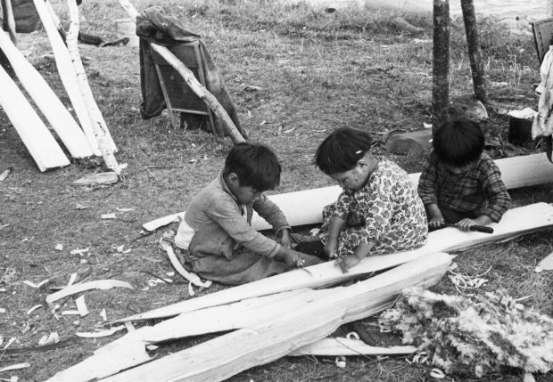 Three young anicinabek who participate in the making of a canoe while sitting on the ground. They are using tools on scrap wood. Black and white picture.