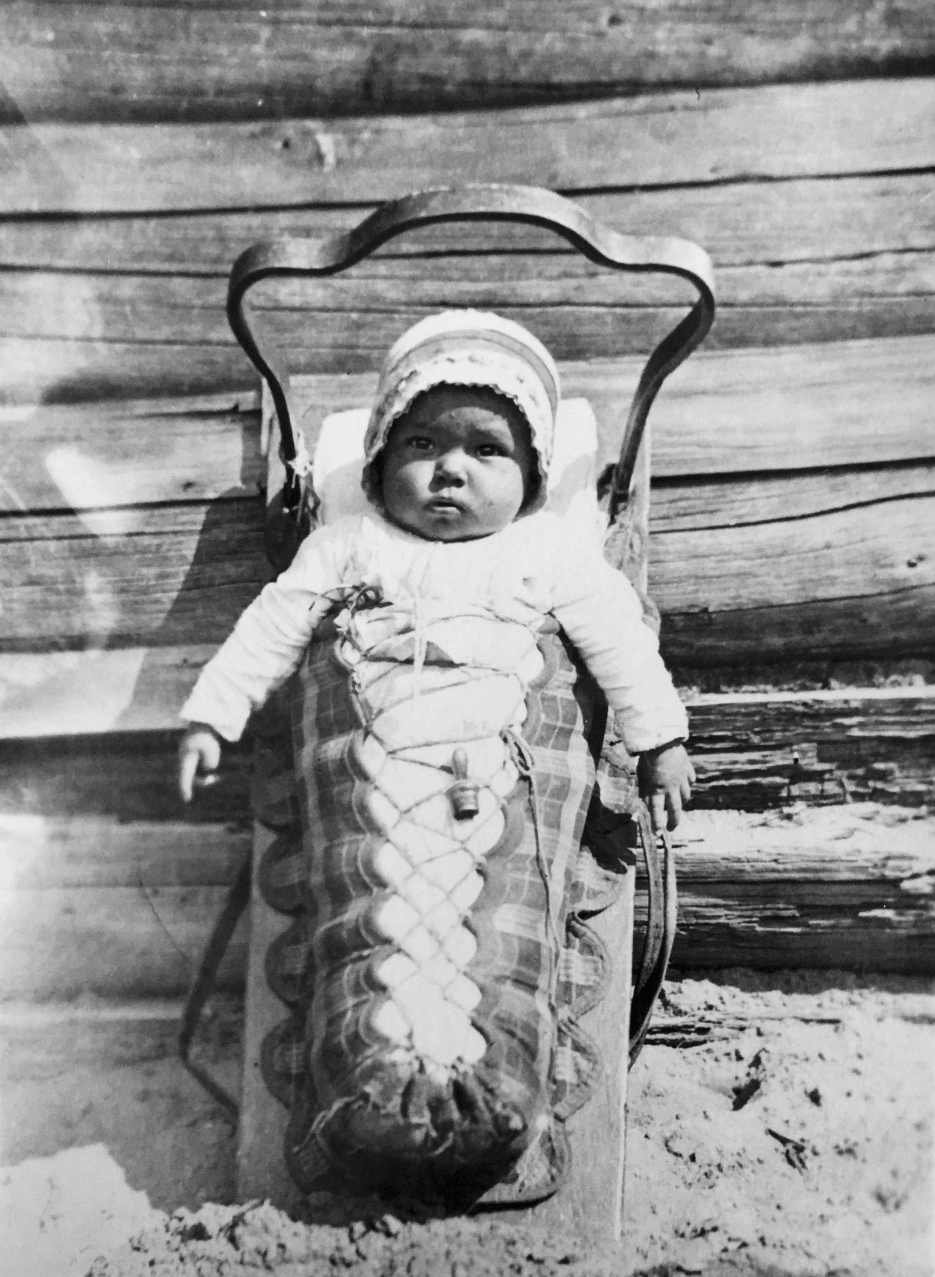 A baby in his baby carrier (tikinagan). He is wearing a hat. His arms are out of the swaddled portion. The baby carrier is placed vertically and is leaning against the wall which allows a baby to see around him. Black and white picture.