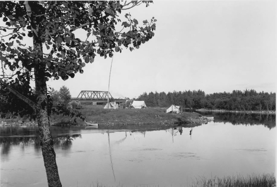 Indigenous camps on an island near Senneterre. We can see some tents and, in the background, a bridge. Black and white picture.