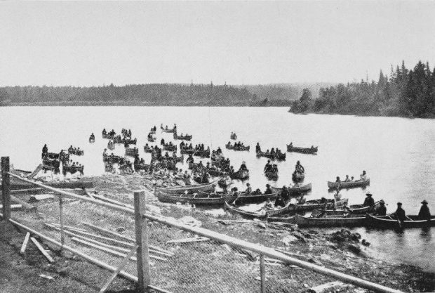 Anicinabek families arrive in Kitcisakik by canoe. We see about twenty canoes in which people are sitting. We see the forest in the background. Black and white picture.