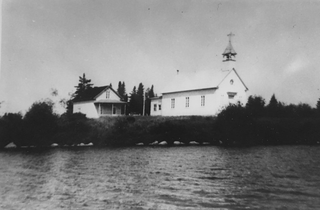 Lac Simon Church as it is located near Lac Simon. We can see another building beside it and the lake it faces. Black and white picture.