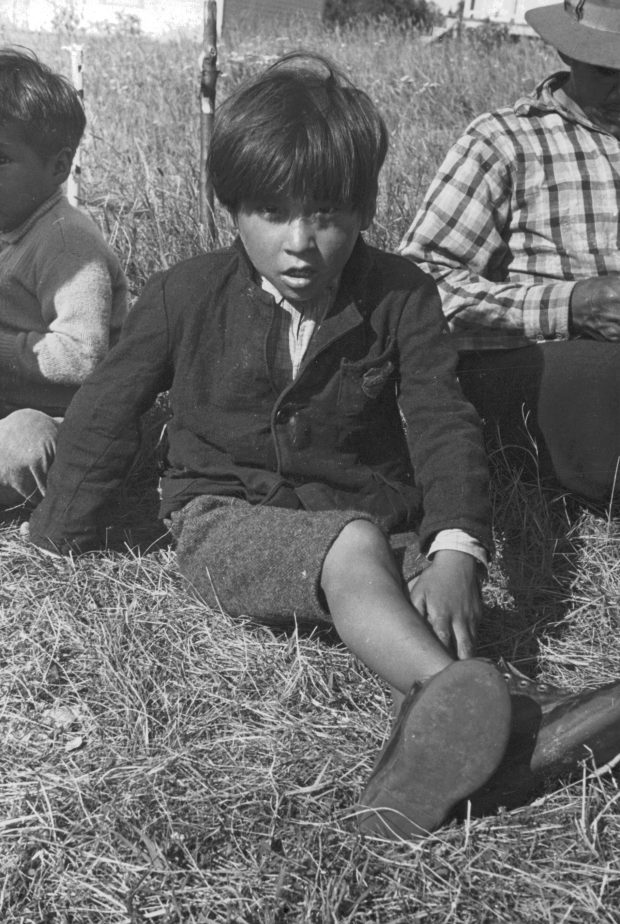 Young Anicinabe boy sitting in the grass near Lake Simon. He is wearing a jacket, shorts, short hair and shoes. A man is on his right and a young man on his left. Black and white picture.