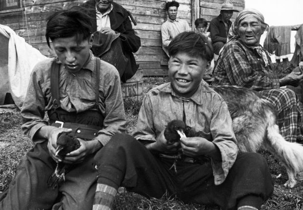 Young anicinabek playing with crows that they hold in their hands. They wear shirts and pants. A few people and a dog surround them. A plank building stands in the background. Fabrics are drying in the wind. Black and white picture.