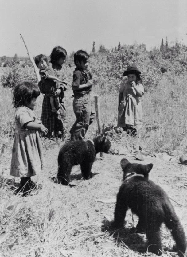 Young anicinabek playing with two small bears outside. Two young girls wearing dresses. Two young boys are standing next to them. One of them is holding a baby in his arms. Around them, we see vegetation. Black and white picture.