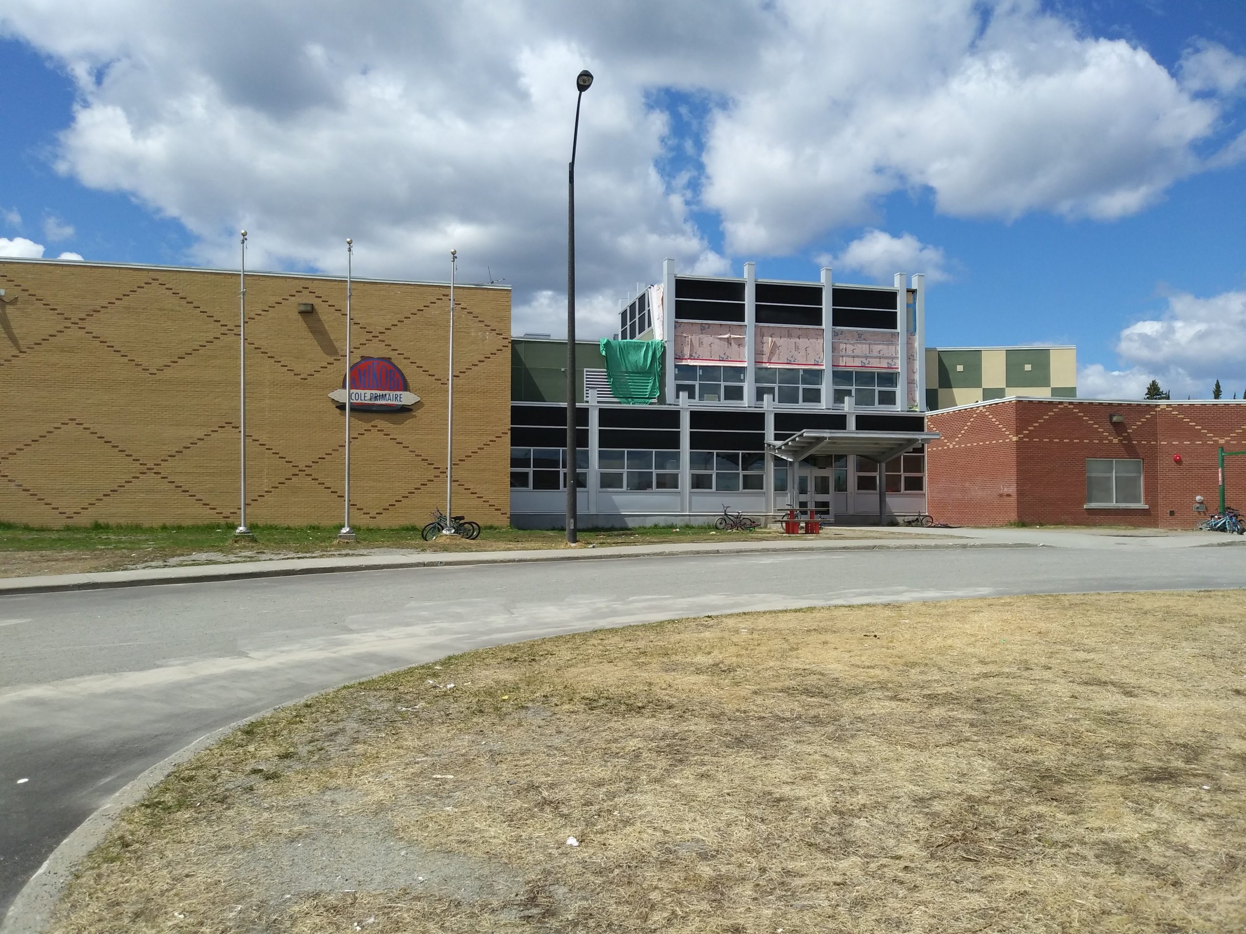 Front of the Lac Simon Elementary School. One portion is made of yellow bricks, the other of red bricks and a windowed portion in the center. We see a portion of the road that leads to the entrance. Picture in color.