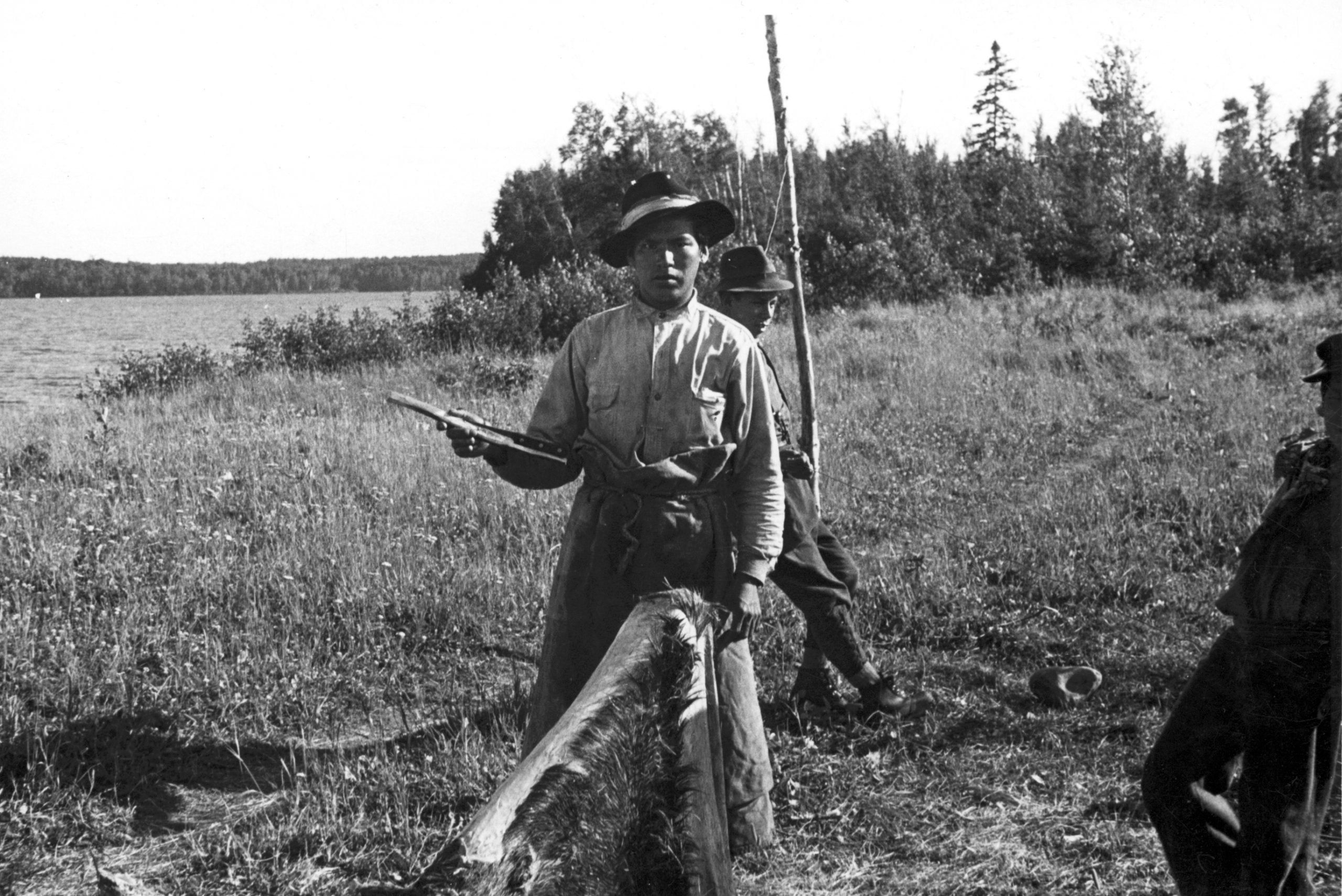 An Anicinabe man holds a traditional scraper in his hand near a hide he is working on. Another man is behind him and one at his side. They are near a lake. Black and white picture.