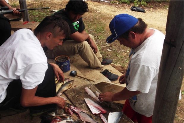 Men watch another man fix a fish. They are sitting on an outside porch of a camp. Picture in color.