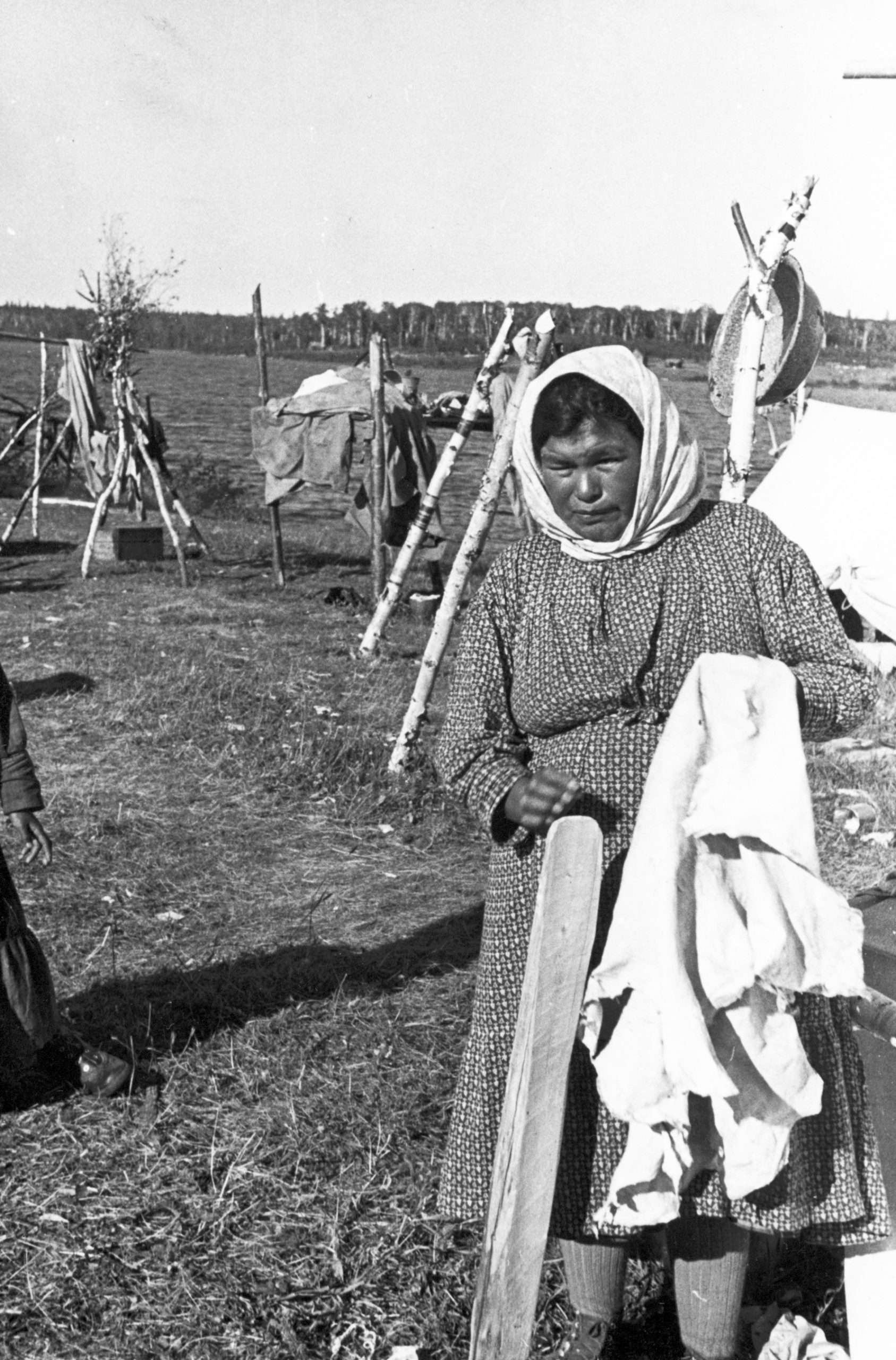 An Anicinabe woman with a tanned skin in her hands. She wears a long dress and a scarf on her head. We see a portion of a camp behind her with tents and wooden supports. Black and white picture.
