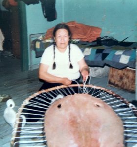An Anicinabe woman sitting on the ground is stretching a beaver skin on a wooden support in the shape of a circle with the help of rope. Behind her, we see a bed. Picture in color.