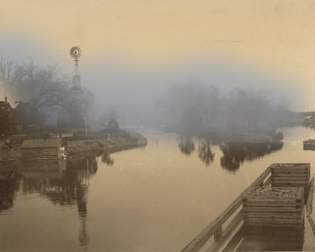 A B&W photo of a canal, with a blue carriage house visible through the mist.
