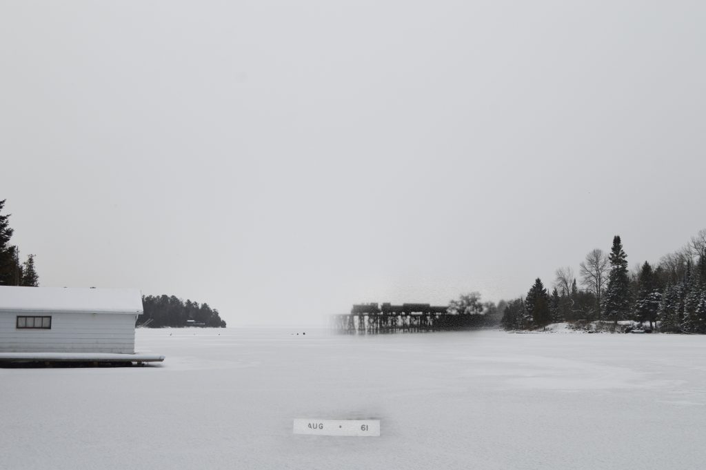 A modern photo of a bay, with a superimposed image of a train crossing a bridge.