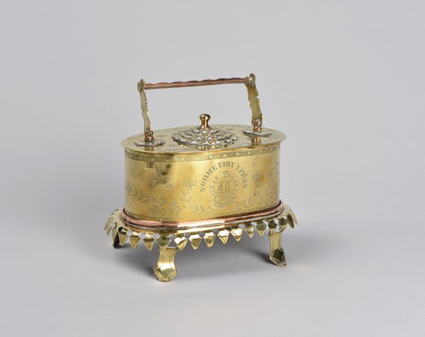 A small, oval-shaped brass jewellery case with a handle and four legs. On the top, there is a small, elaborately decorated combination lock.