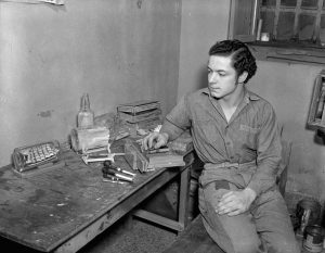 German prisoner of war is sitting at a workbench and is looking at the ship in a bottle he has made.