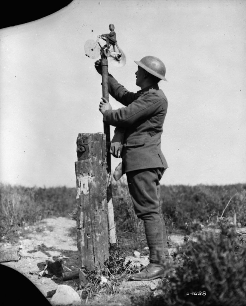 A First World War soldier is installing a weather vane on the top end of a metal pipe attached to a post. The vane is a sculpture of a man riding a bike.