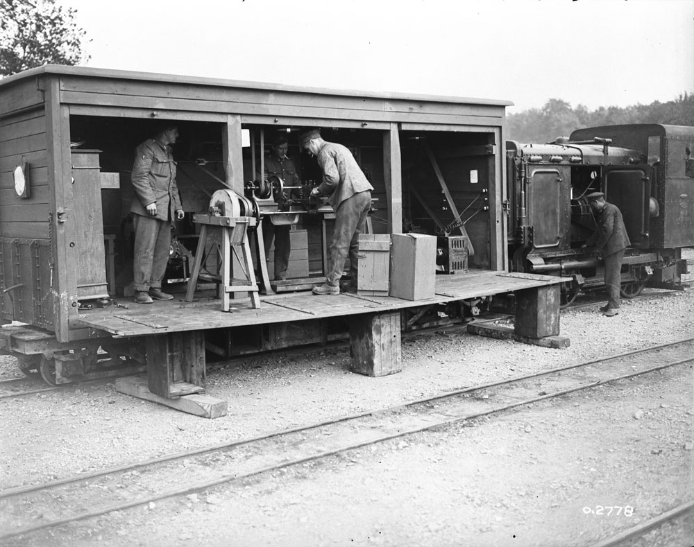 Photo en noir et blanc d’un atelier ambulant logé dans un wagon de tramway en bois. Un côté est ouvert et laisse voir trois soldats occupés à fabriquer divers articles.