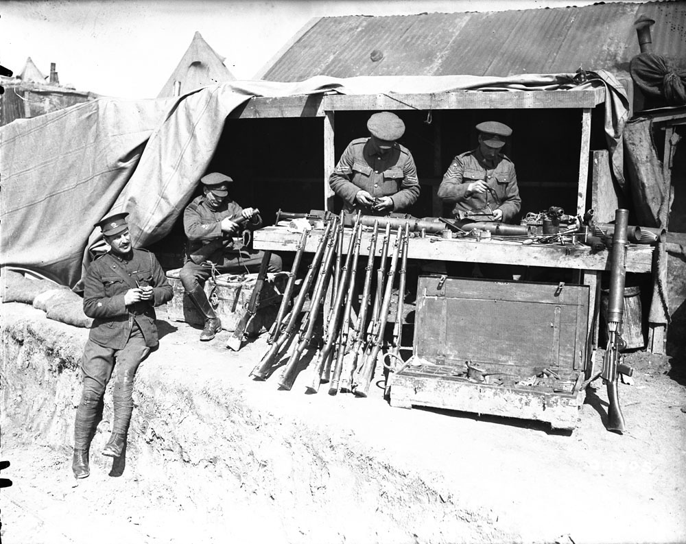 A black and white image of an impromptu armourer’s shop. There are two soldiers behind a table displaying various weapons, giving attention to their goods while two other soldiers are present.