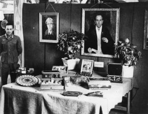 Prisonnier de guerre allemand en uniforme près d’une table couverte d’objets d’artisanat.  Ses mains sont derrière le dos et il a un sourire moqueur.