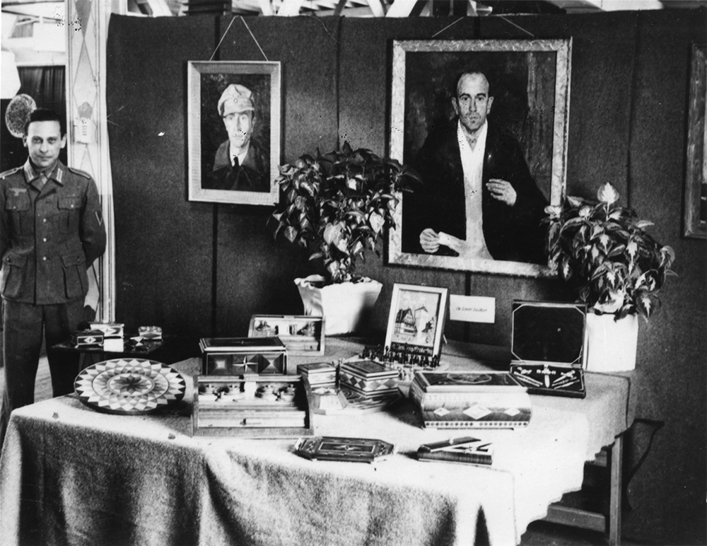 A uniformed German prisoner of war is standing behind a table covered in handmade crafts. He has his hands behind his back and he is grinning at the camera.