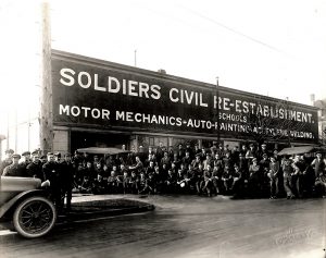 Photo en noir et blanc d’un groupe important d’anciens combattants, vêtus en civil, posant à l’extérieur, devant leur atelier.
