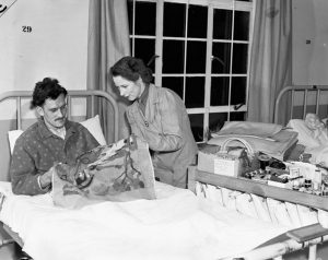 Photo en noir et blanc d’un soldat de la Seconde Guerre mondiale, en pyjama et couché sur un lit d’hôpital. Une travailleuse de la Croix-Rouge canadienne lui montre  comment effectuer une broderie.