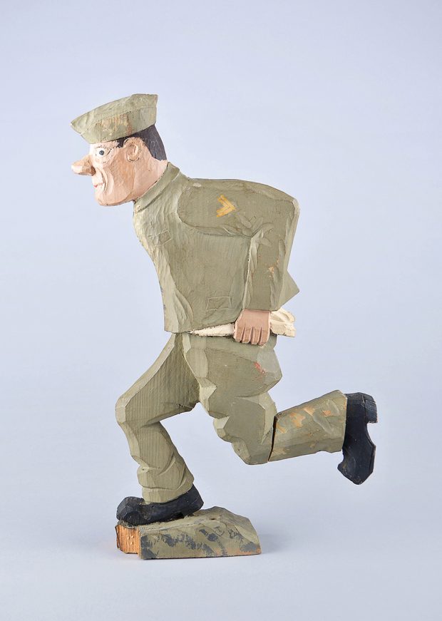 A rustic painted wooden carving of a soldier running. He has a green uniform and black shoes and his shirt is untucked.