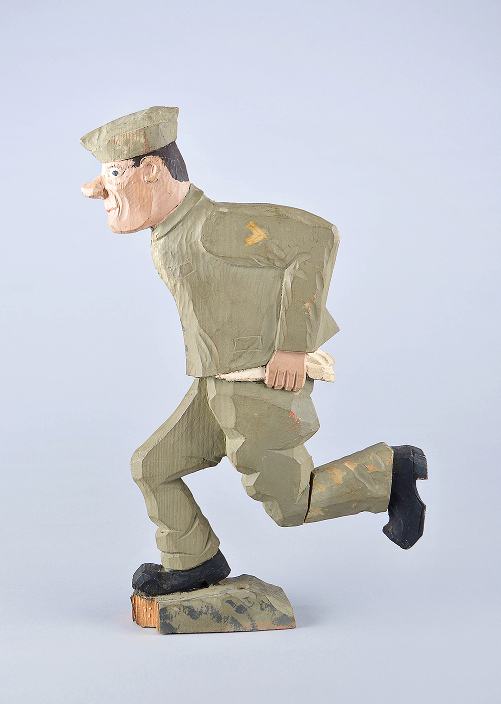 A rustic painted wooden carving of a soldier running. He has a green uniform and black shoes and his shirt is untucked.