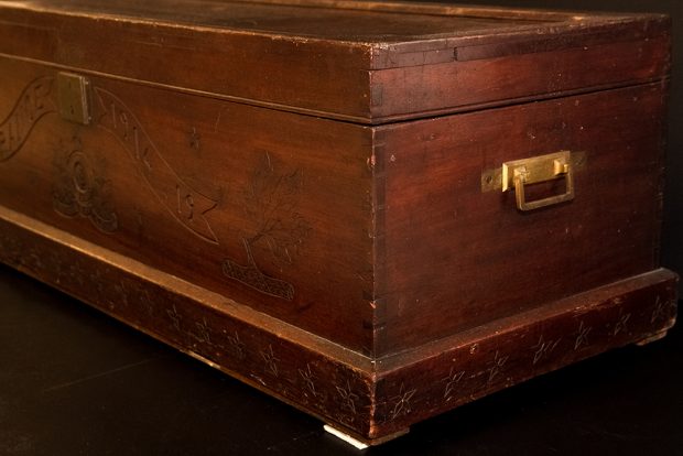 The corner of a handmade wooden footlocker showing engraving and a brass handle.