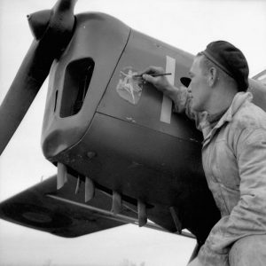 A black and white photo is touching up an illustration he is painting on the nose of an airplane.