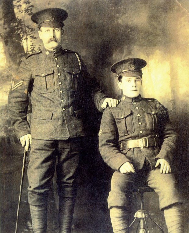 An old photograph of two serious soldiers. One is standing up and leaning on a cane with his hand on the shoulder of the other soldier.