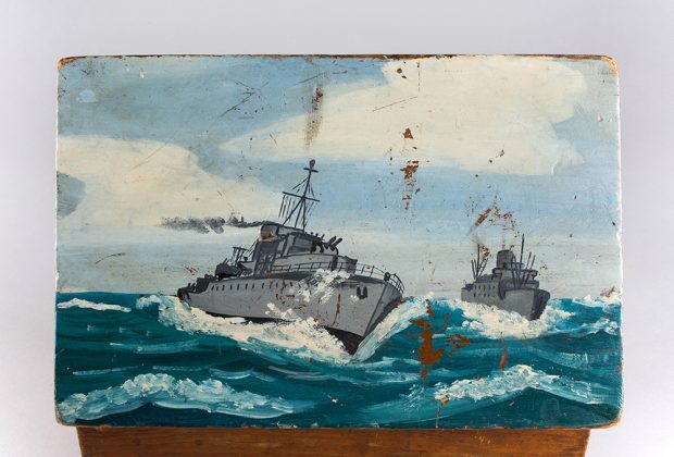 A close-up picture of the top of a wooden ditty box. It is painted with the image of two naval ships on the stormy seas.