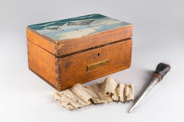 A wooden ditty box with ships at sea painted on the lid. In front of it is a strip of canvas with needles pierced through and well-worn, makeshift awl.