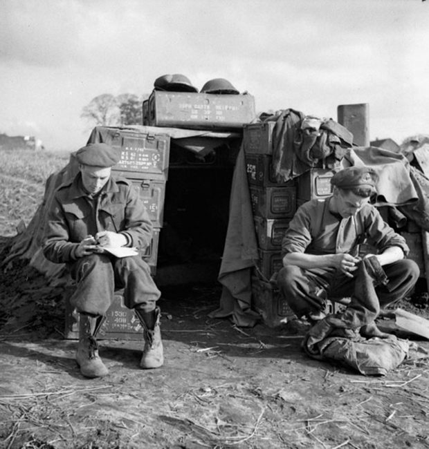 Black and white photo of two Bombardiers sewing in front of a makeshift house in on the edge of a field.