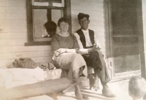 In this sepia-coloured photograph, a young smiling couple are sitting close together. They have their legs crossed and are sitting on a porch and looking at the camera.