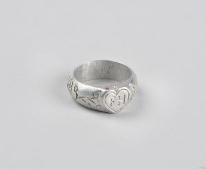 An aluminum ring engraved with plants around the outside of the ring. There is a heart filled with the initials “F H” on the top.