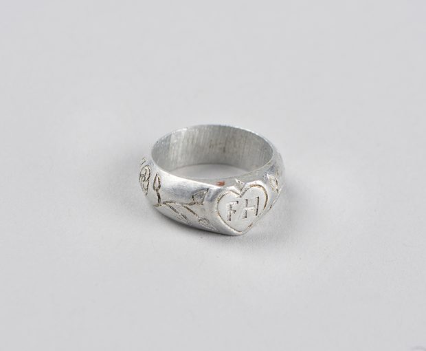 An aluminum ring engraved with plants around the outside of the ring. There is a heart filled with the initials “F H” on the top.