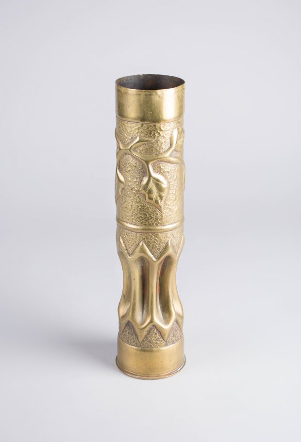 A brass vase shaped from a shell casing. It is indented at the bottom and embossed with an ivy with leaves around the top.