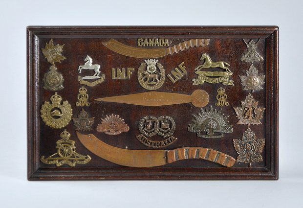A wooden frame around a wooden panel covered in various metal badges and three metal letter openers.