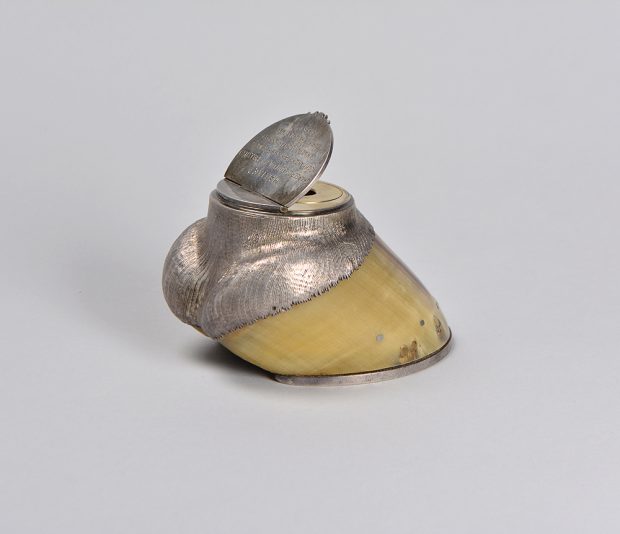 A horse’s hoof with finished silver forms at both ends. The silver is shaped like the lowest part of the horse’s leg at the top of the hoof and has a lid opening into an ink well.