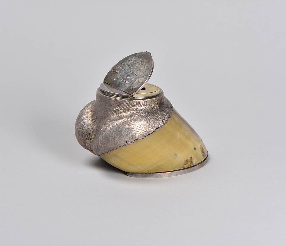 A horse’s hoof with finished silver forms at both ends. The silver is shaped like the lowest part of the horse’s leg at the top of the hoof and has a lid opening into an ink well.