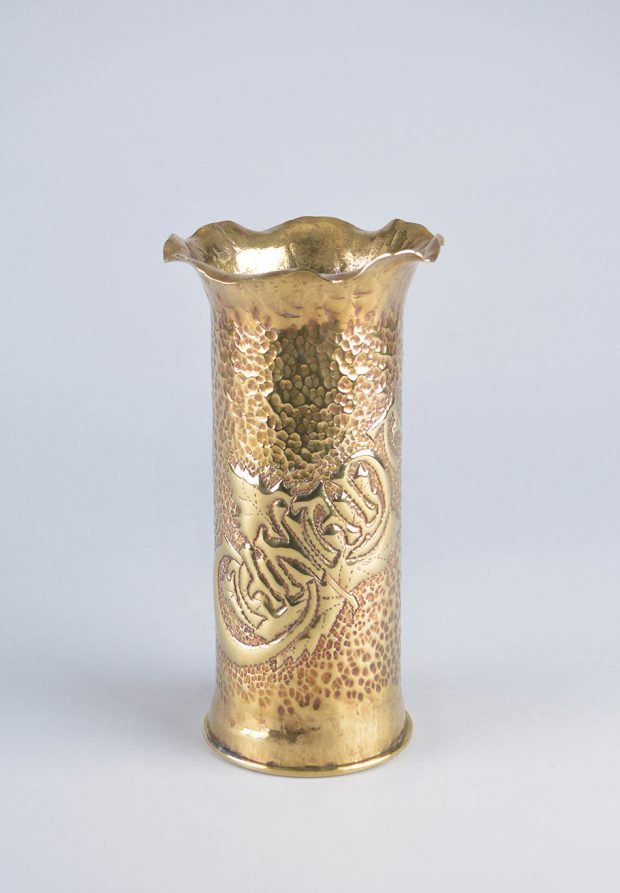 Tall cylinder brass vase with ribbing on the bottom and a rippled opening. It is covered in patterned dinting and engraved with the word “Canada” at an upward angle.
