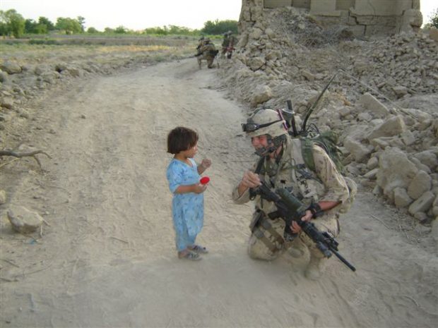 A color photo of a kneeling soldier in full combat uniform given a very young child a knitted Izzy Doll.