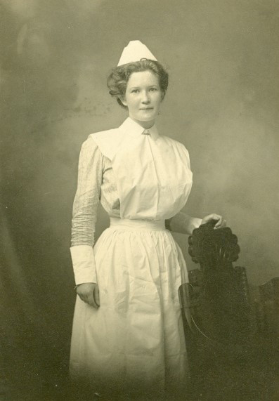 A sepia picture of a nurse in full uniform with her hand on the back of a tall carved wooden chair.