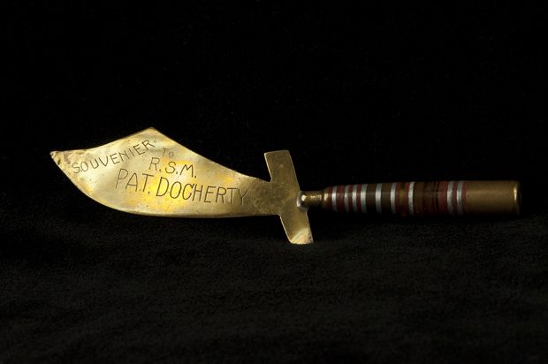 A colour image of a letter opener shaped like a saber. The handle, made from a bullet, it attached to a blade made of polished brass and is engraved.