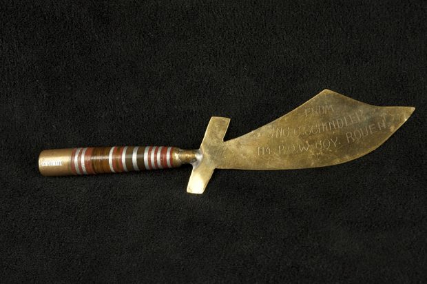 A colour image of a letter opener shaped like a saber. The handle, made from a bullet, is attached to a blade made of polished brass and is engraved.
